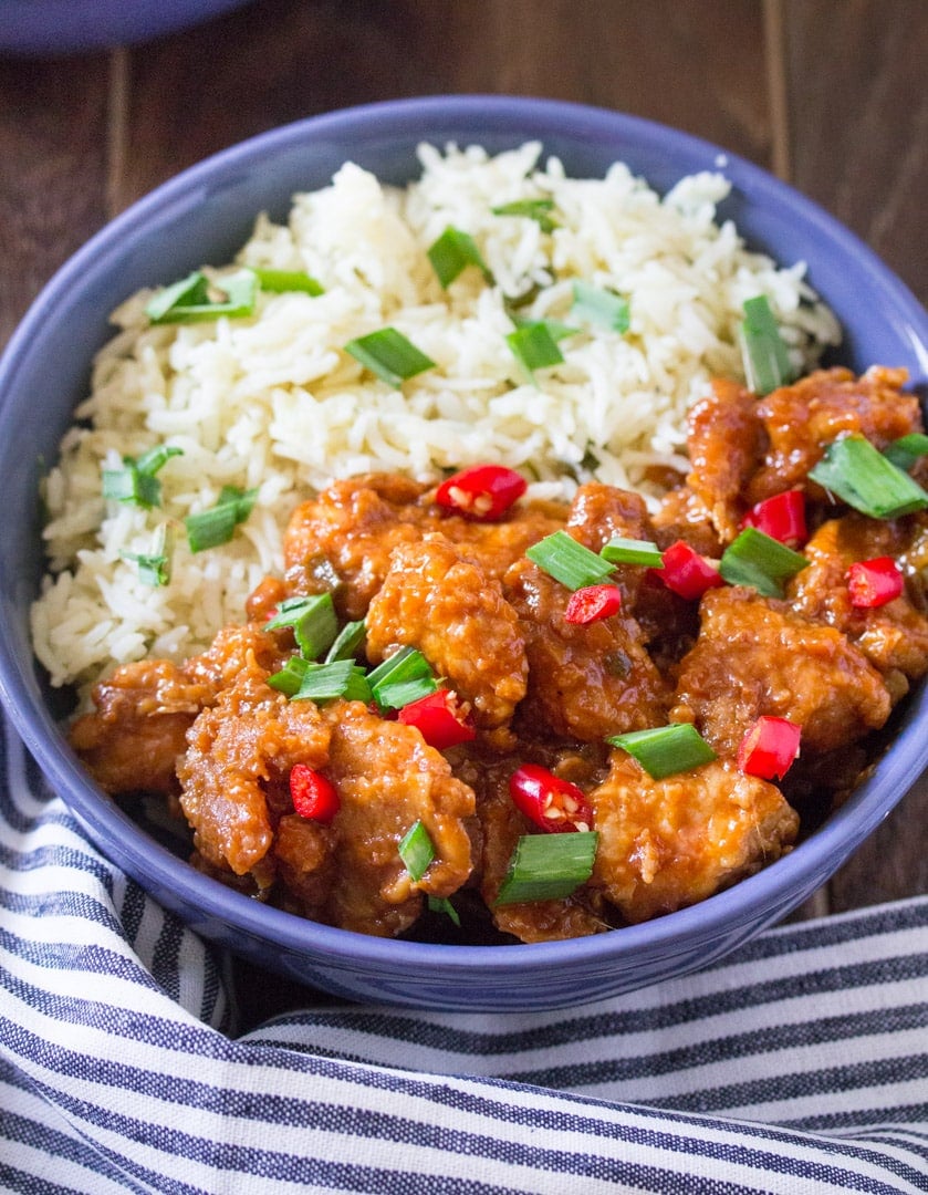 overhead shot of chicken manchurian in gravy with white rice and green onions in a blue bowl with a blue and white striped towel