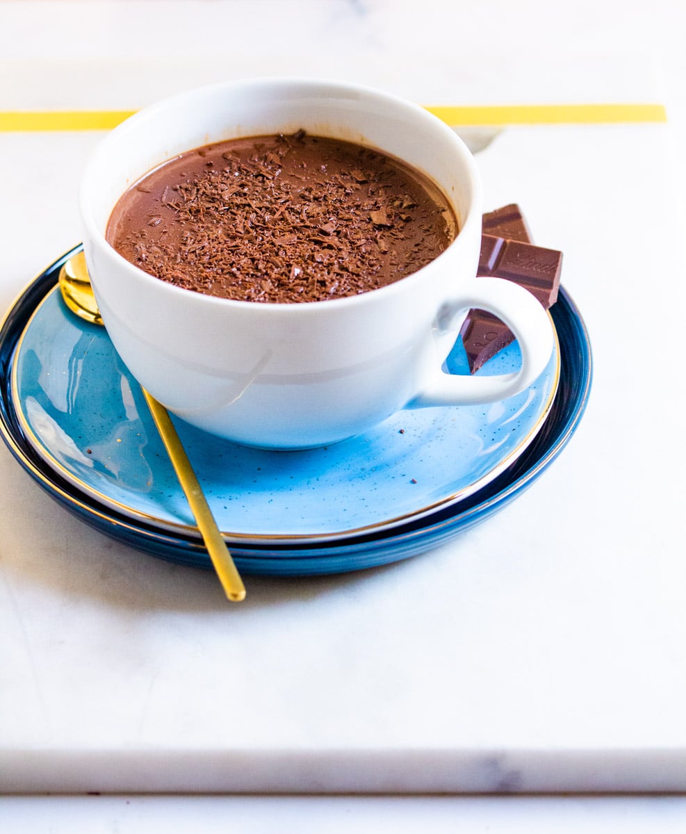 Cup of hot chocolate on a blue saucer with chocolate shavings on top and chocolate pieces on the plate.