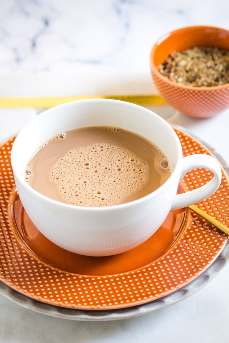 Creamy chai in a white mug on an orange plate with bowl of spices in the background.