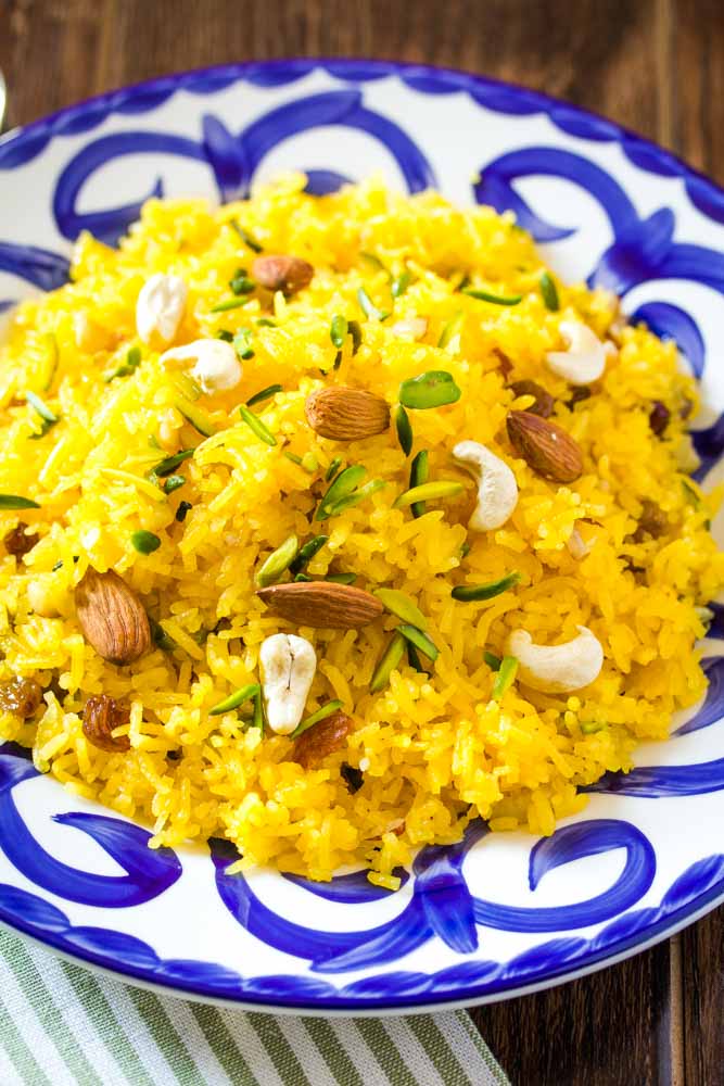 A close up shot of meethe chawal served on a blue and white patterened plate, topped with nuts. 