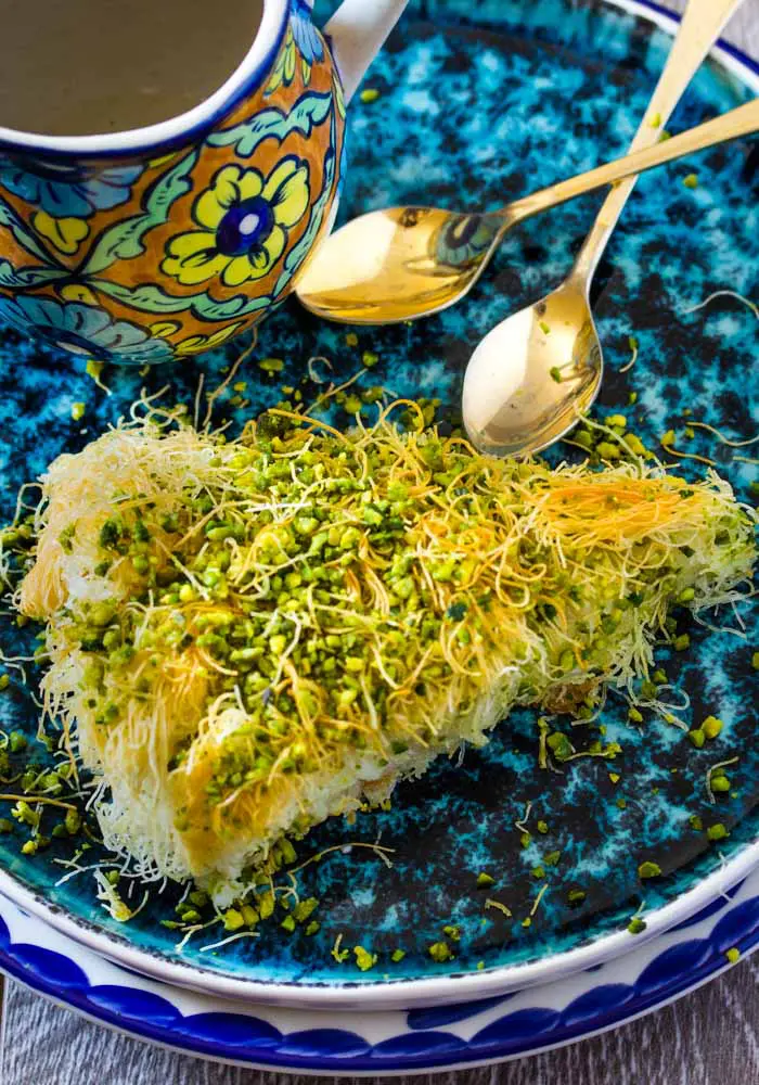Slice of Kunafa on a turquoise plate next to two gold spoons.