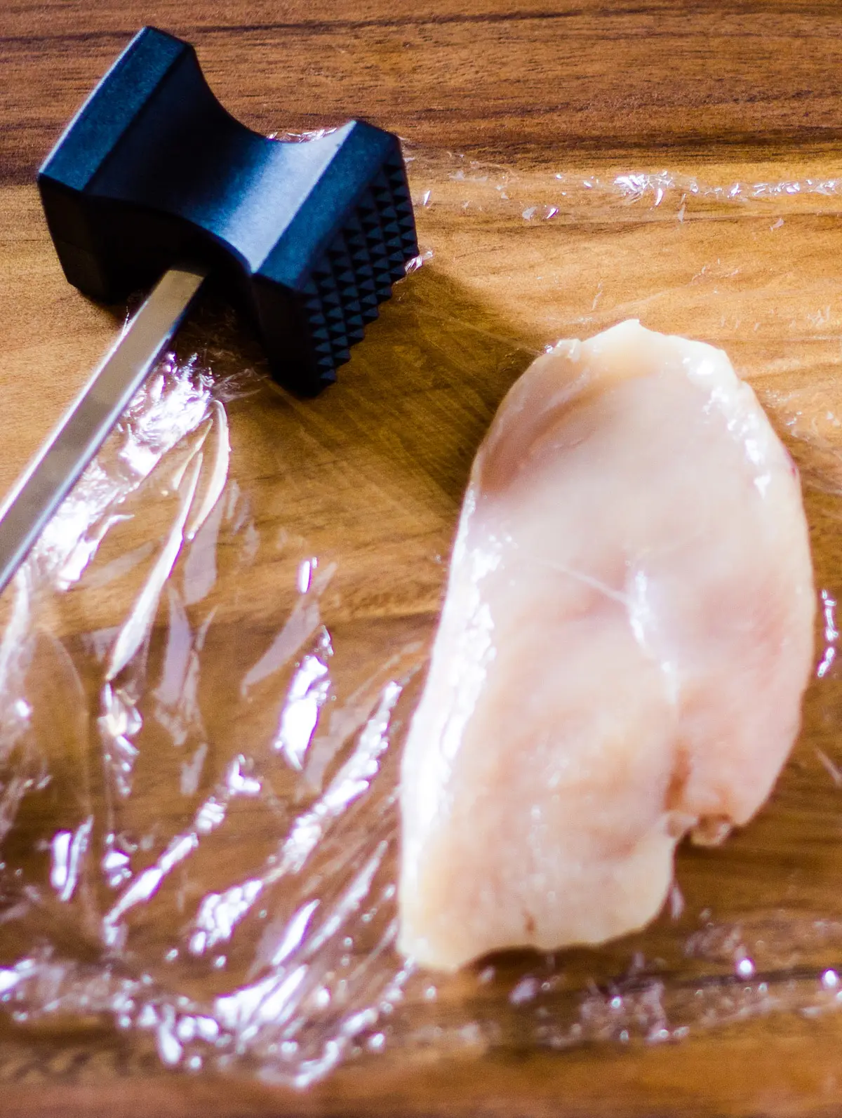 A chicken breast and a kitchen mallet on a wooden cutting board.