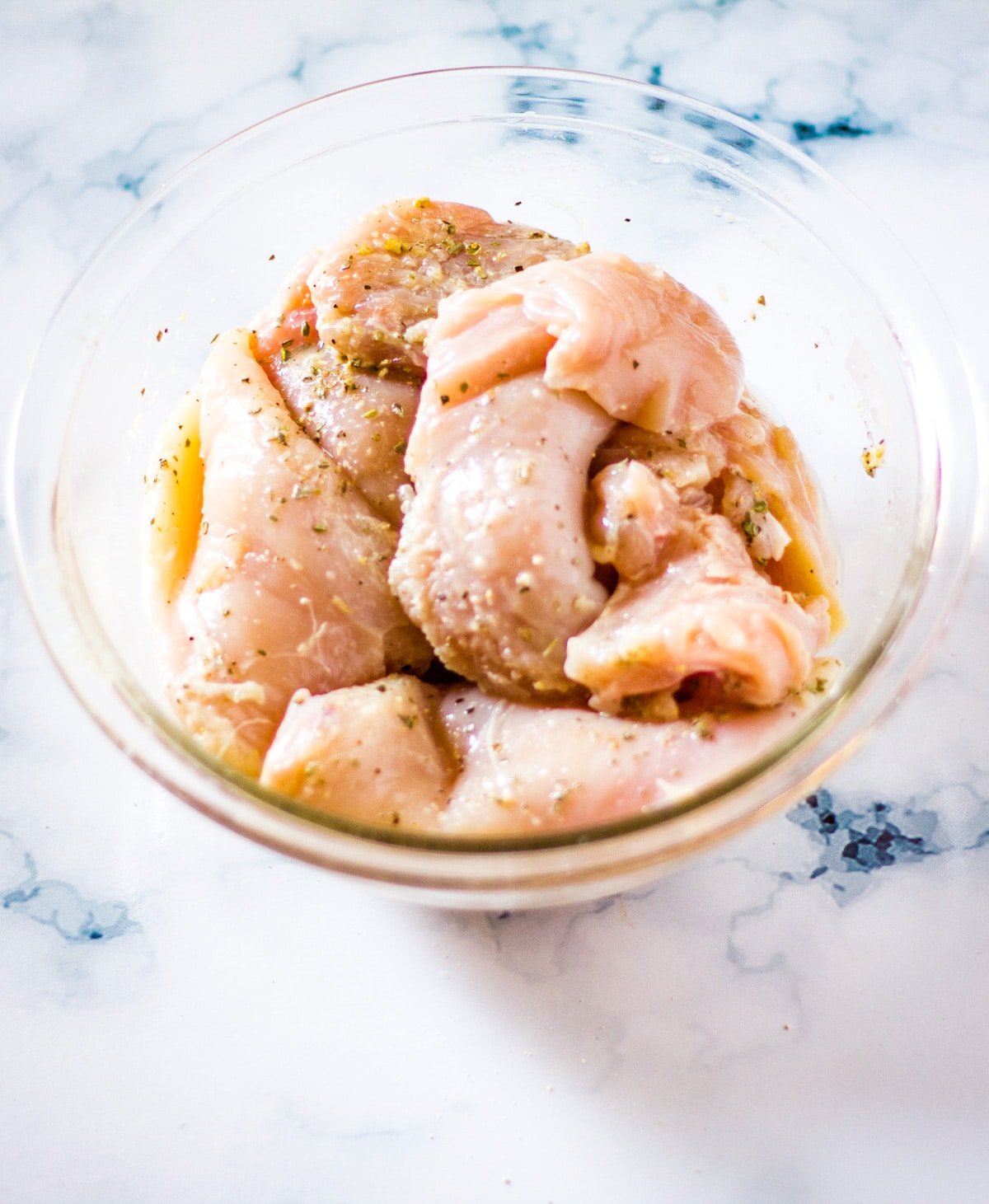 Marinated chicken in a glass mixing bowl on a white marble backdrop.