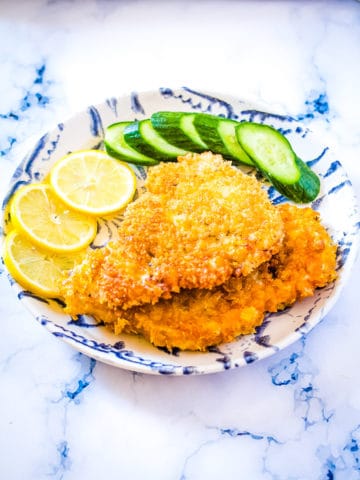 Breaded chicken cutlets on a blue and white plate with lemon slices and sliced cucumber.