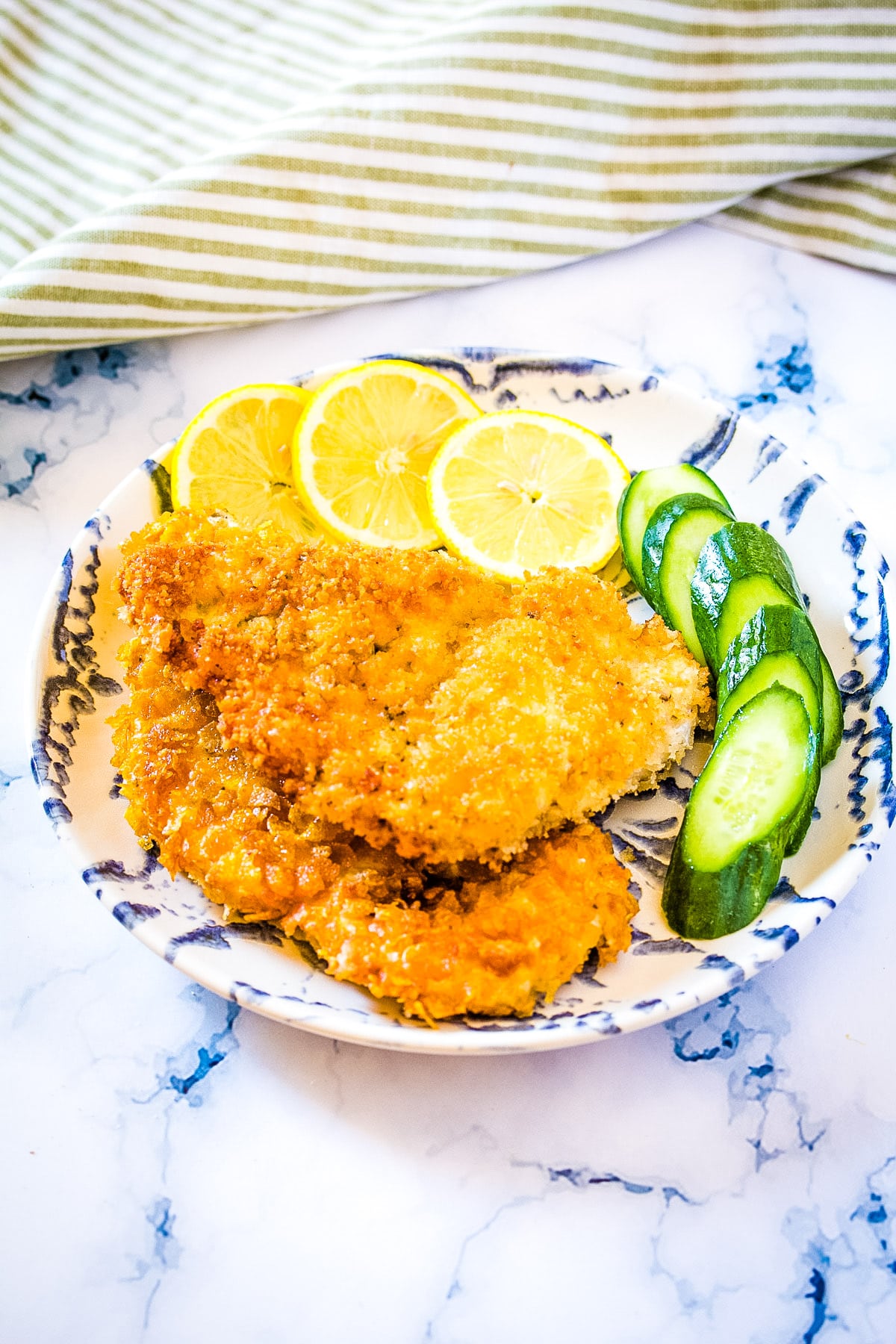 Two crispy chicken cutlets on a blue and white plate with sliced lemon and sliced cucumber.