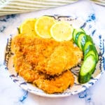 Breaded chicken cutlets on a blue and white plate with lemon slices and sliced cucumber.