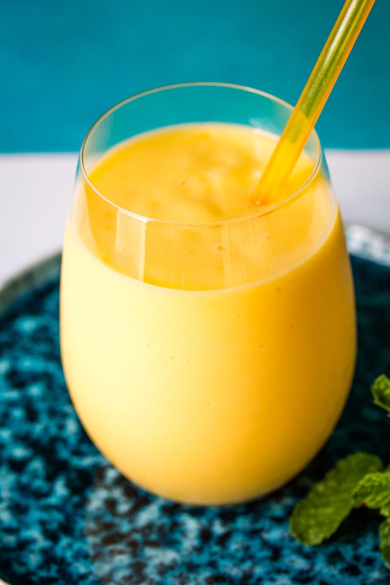 Bright yellow mango milkshake in a clear glass on a blue plate. 