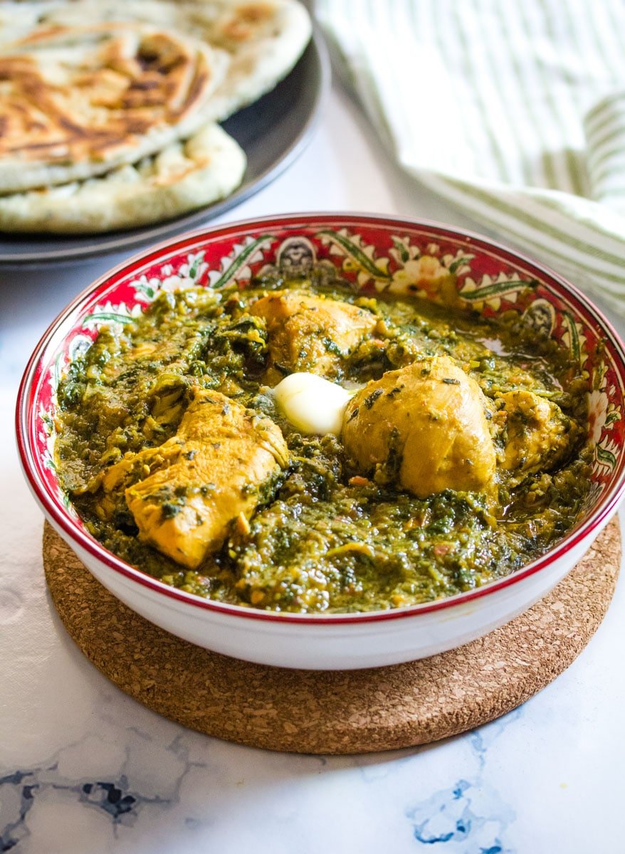 Palak chicken topped with butter in a red bowl and naan bread on a side dish.