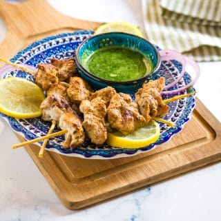 skewers of golden brown malai tikka place on a blue plate next to a bowl of green chutney.