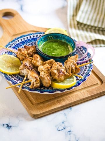 skewers of golden brown malai tikka place on a blue plate next to a bowl of green chutney.