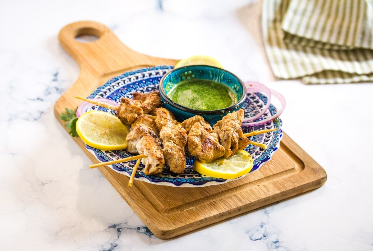 a blue plate of grilled chicken skewers placed on a small wooden cutting board.