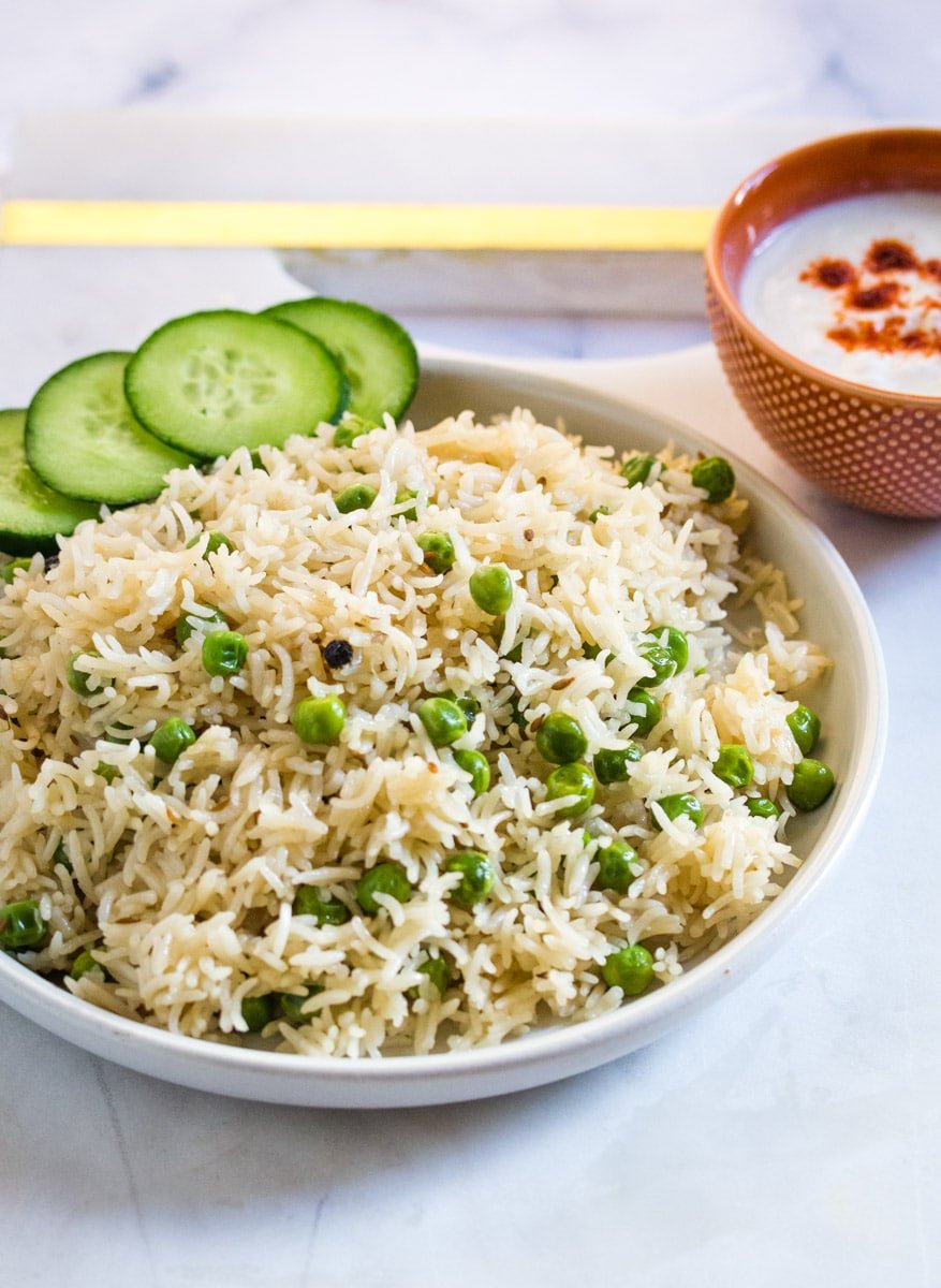 Matar pulao served with sliced cucumbers and a bowl of yogurt raita.