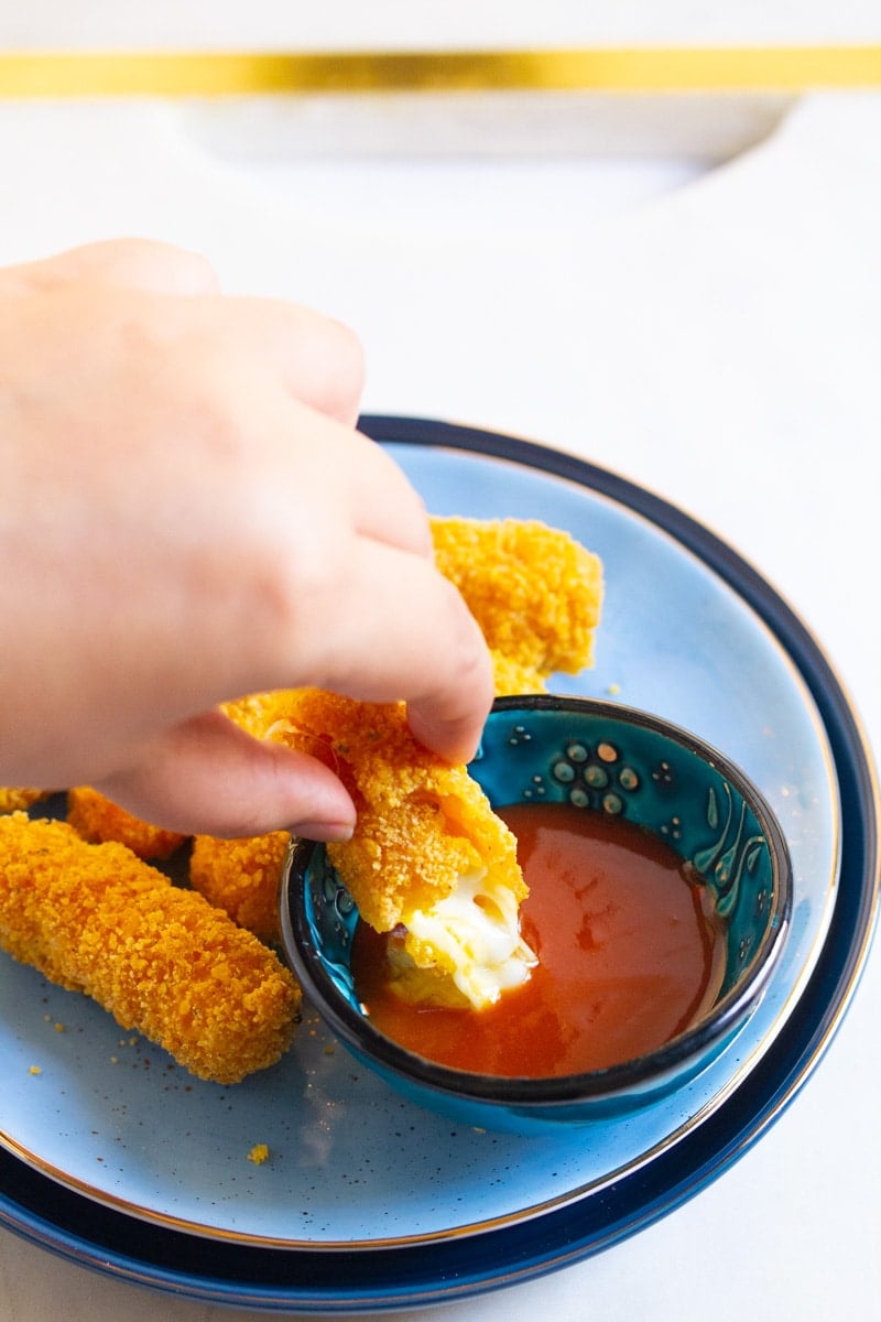 A hand dipping Doritos mozzarella stick into sriracha sauce.