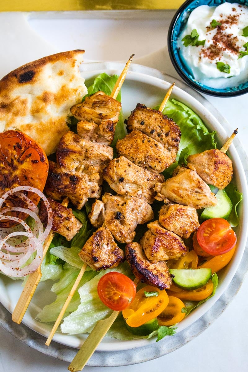 Three skewers of chicken sheesh tawook laid on a bed of green lettuce and served with salad.