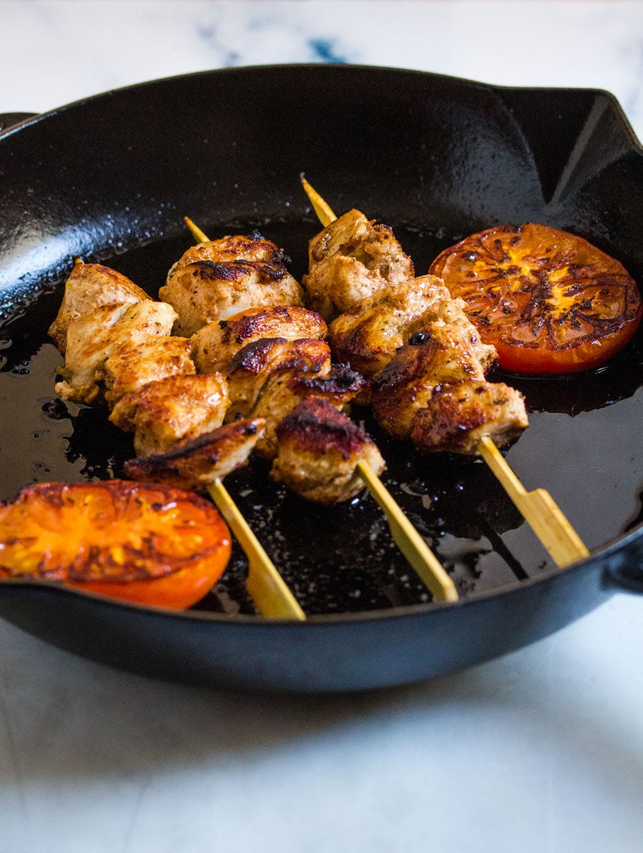 Three skewers of grilled chicken in a black cast iron pan with grilled tomatoes.