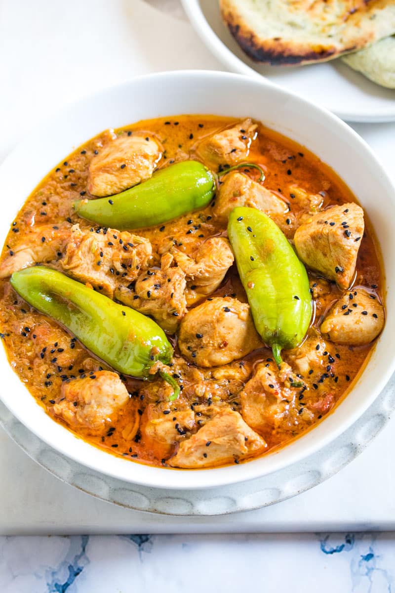 Achari Chicken served in a white bowl, topped with three big green chilies.