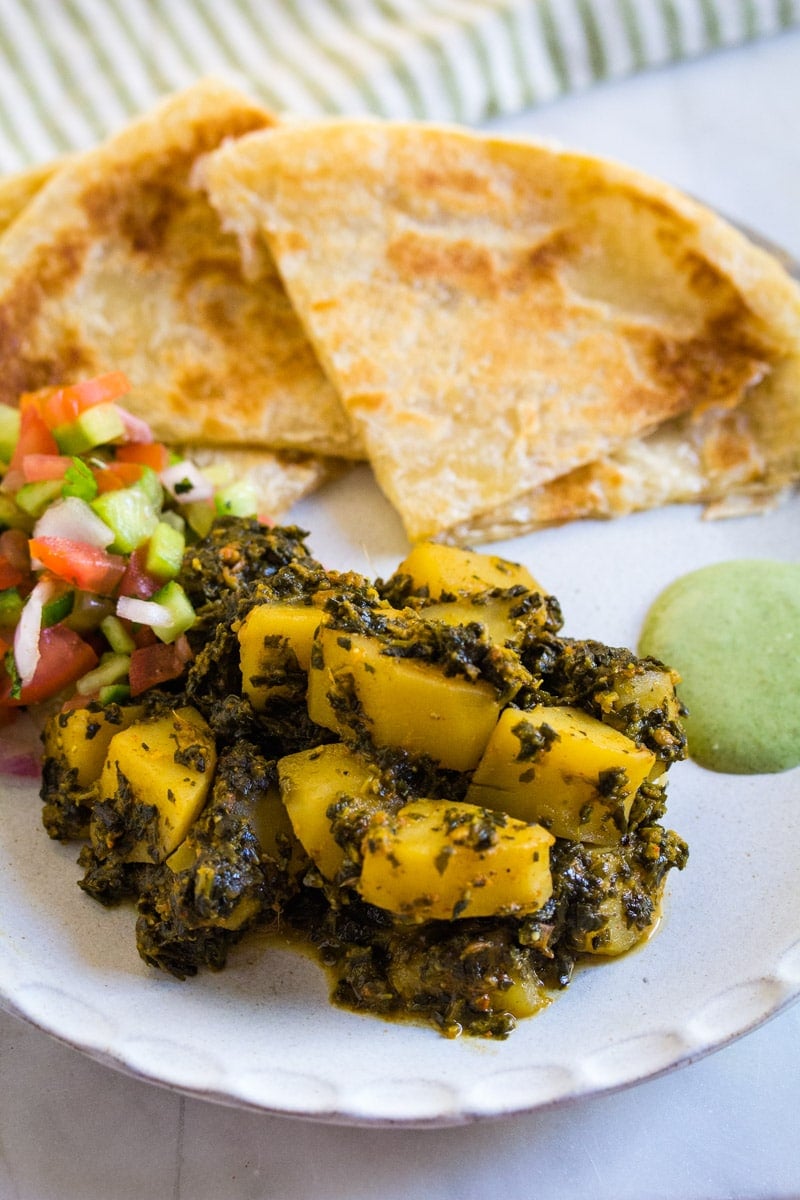 A serving of spinach and potato curry with salad, mint raita and paratha on a white plate.