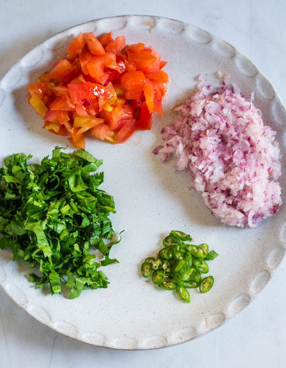 Chopped tomatoes, onions, cilantro, and green chilies laid out on a light grey plate.