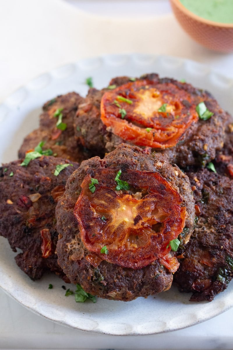 A close up shot of four chapli kababs on a grey plate, served with a bowl of mint raita for dipping.