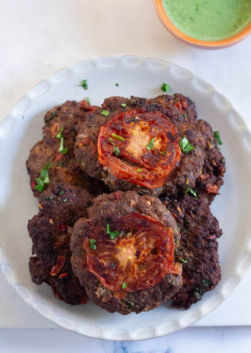 An overhead shot of chapli kebab topped with tomato slices and a side of mint raita.