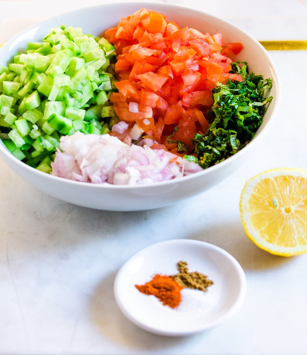 A bowl of chopped vegetables, half a lemon and a small plate of spices laid out on a white marble slab.