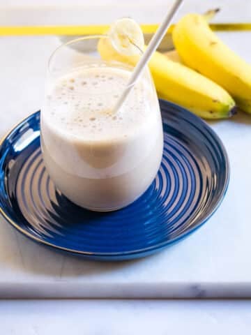 Banana milk served in a glass on a blue plate, with two bananas in the background.