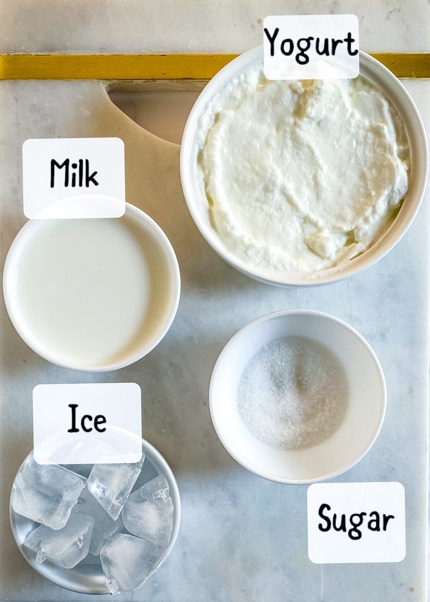 Ingredients of sweet lassi laid out on a white marble slab.