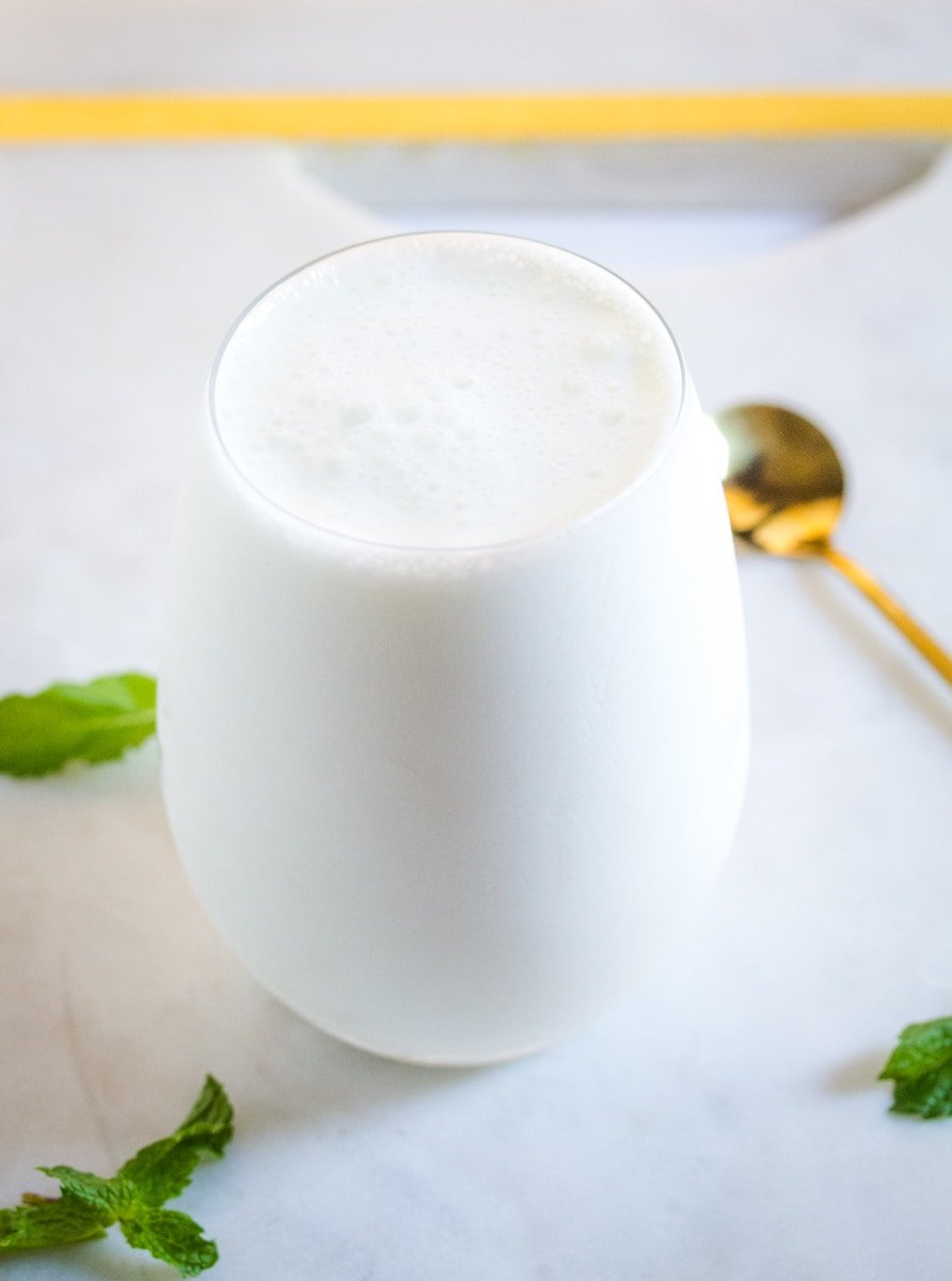 A glass of an Indian yogurt drink with a golden spoon and fresh mint leaves on a white marble slab.