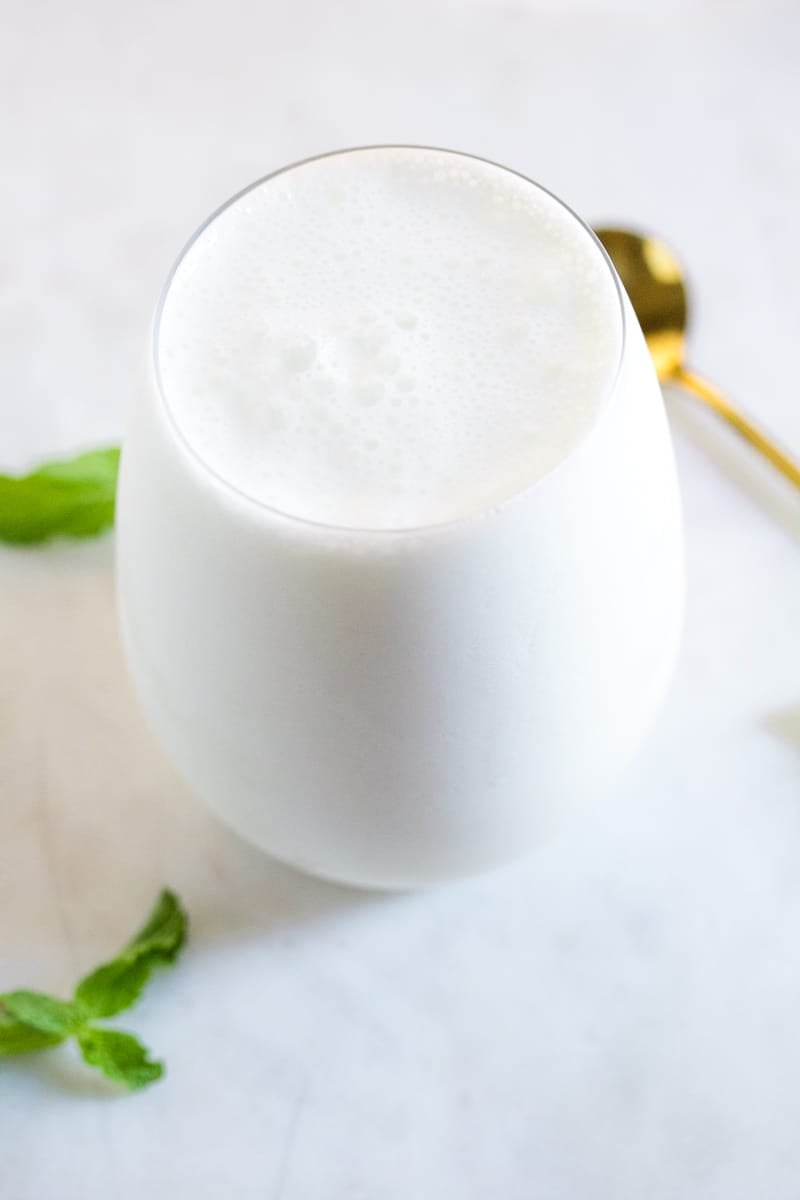 A glass of sweet lassi placed on a white marble slab, next to a gold spoon and two mint leaves.