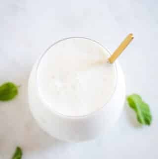 Overhead shot of meethi lassi served in a round glass with a golden spoon, on a white marble slab.