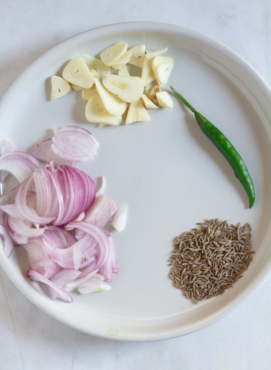 Tempering ingredients: sliced red onion, cumin seeds, green chili, and garlic laid out on a light grey plate.