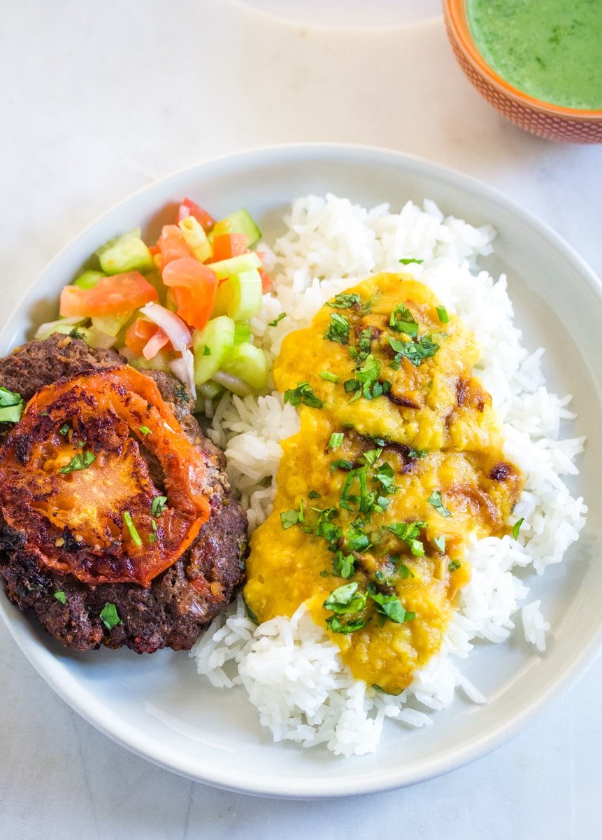 A plate of red lentils served with white jasmine rice, chapli kabab, and kachumber salad.