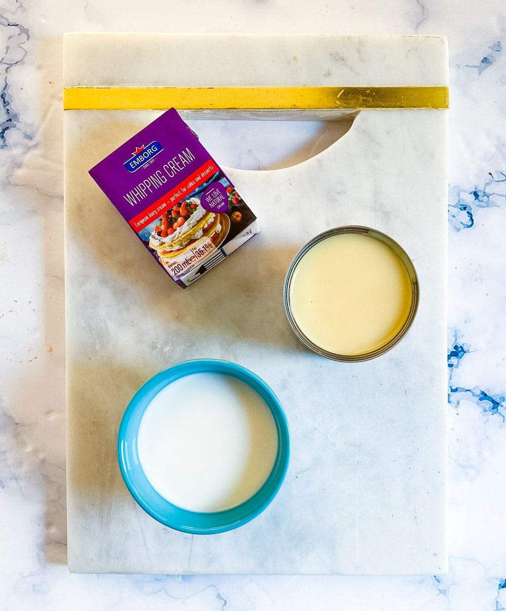 Whipping cream, a bowl of milk, and a can of sweetened condensed milk placed on a white marble slab.