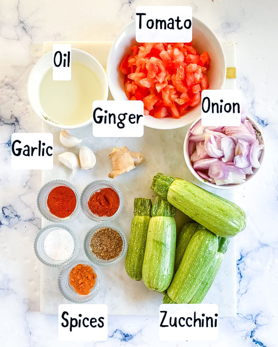 Ingredients to make zucchini curry laid out on a white marble backdrop.