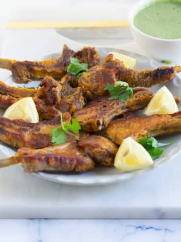 A plate of Pakistani mutton chaap fry garnished with lemon wedges and cilantro leaves.