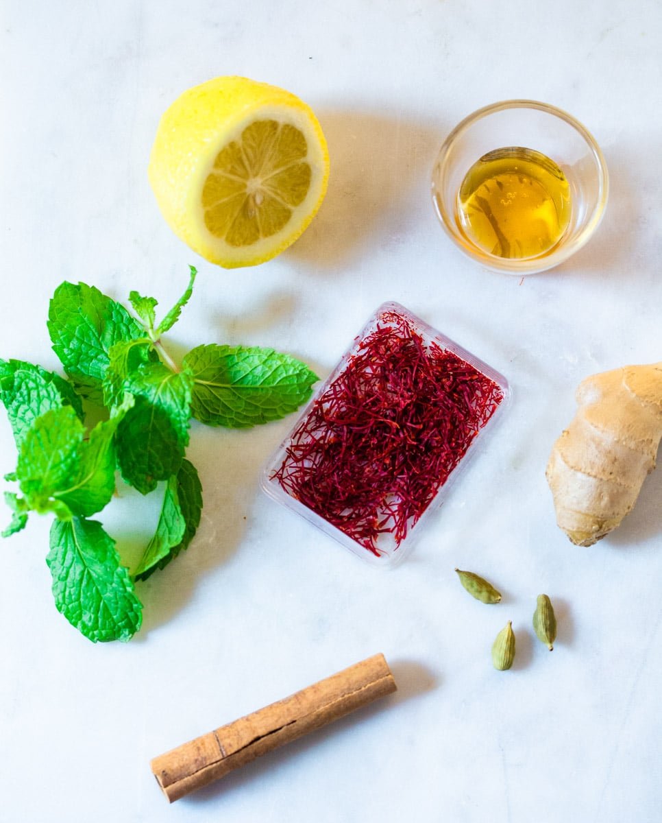 A pictures of ingredients needed to make saffron tea: lemon, saffron threads, mint, cinnamon, cardamom, honey, and ginger.