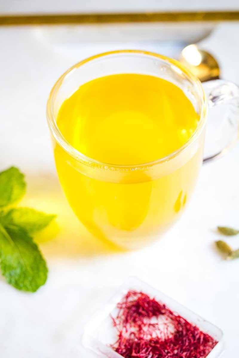 Saffron Tea served in a clear glass mug, surrounded by fresh mint leaves, cardamom, and saffron threads.