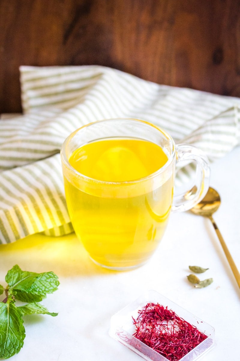 A clear mug of saffron tea, placed on top of a white marble slab that is scattered with saffron threads, mint leaves, and cardamom pods.