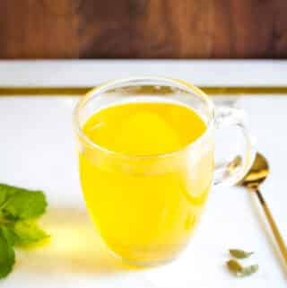 A clear glass mug of saffron tea, surrounded by mint leaves, a box of saffron, and cardamom.