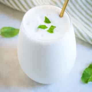 Salty lassi is served in a clear glass goblet, and placed on top of a white marble slab that has three mint leaves scattered on it.